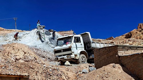 A lorry unloads gravel at a construction site in a mountainous desert landscape under a clear sky,