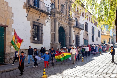People march in a parade through a sunny street with historic buildings and flags, demonstration in