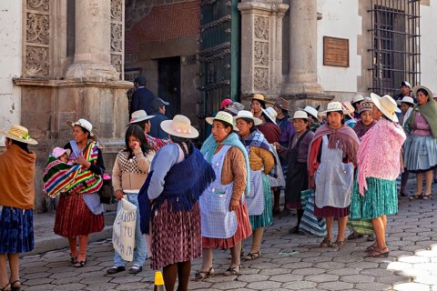 Women in traditional dress stand on a busy cobbled street in front of a historic building,