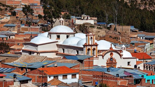 A city view with domes of a church in the foreground, surrounded by brick houses in a mountainous