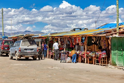 Various market stalls with visitors and cars, lively rural atmosphere under a cloudy sky, market