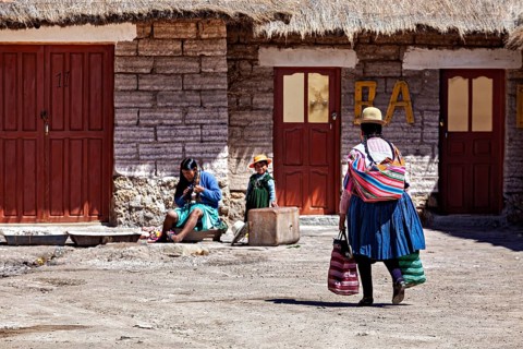 A woman in traditional dress carries bags while children and another person sit in the background,