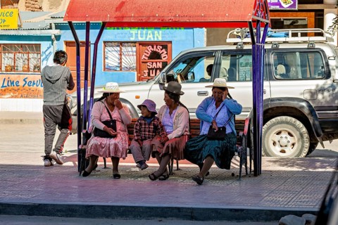 People in traditional dress sit on a bench in an urban environment as a car drives past, people at