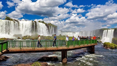 People walk on a bridge in front of an impressive waterfall under a blue sky, The Iguazu Falls