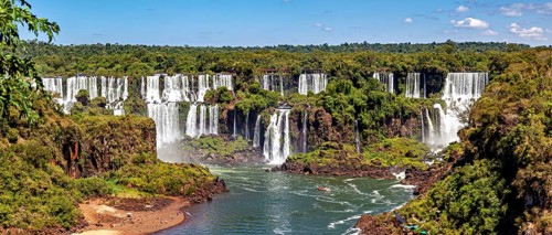 An impressive panoramic view of a cascade of waterfalls flanked by lush greenery and a clear sky,