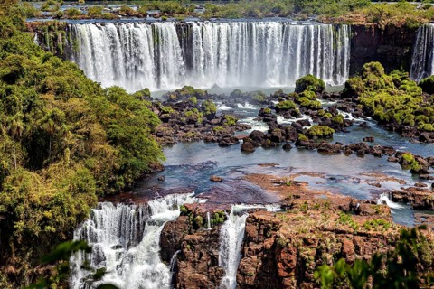 Waterfalls cascade over rocks into a wide river surrounded by dense forest, The Iguazu Falls
