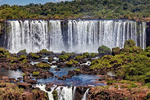 Wide waterfalls cascade over rocks into a river surrounded by lush greenery, The Iguazu Falls