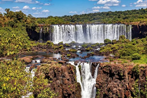 Steep waterfalls cascade over rocks, surrounded by dense greenery and a cloudy sky, The Iguazu