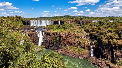 Large waterfalls flow over steep cliffs, surrounded by lush greenery and a blue sky with clouds,