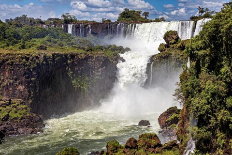 Large waterfall with dense spray and surrounding green forests under a blue sky, The Iguazu Falls