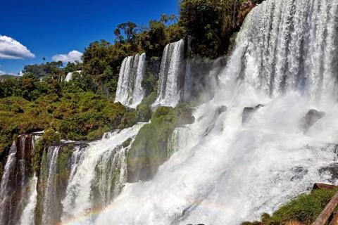 Massive waterfalls create dense spray and a rainbow, surrounded by green forest and blue sky, The