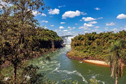 A wide river flows through a green landscape with jungle, while a distant waterfall adorns the