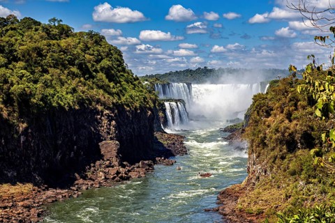 Majestic waterfalls flow between lush vegetation and steep rock faces under a clear sky with white