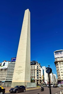 A tall obelisk under a clear blue sky surrounded by modern buildings, The Great Obelisk in the city