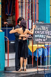 A couple dances tango in front of a colourful building, hugging each other, next to a menu board,