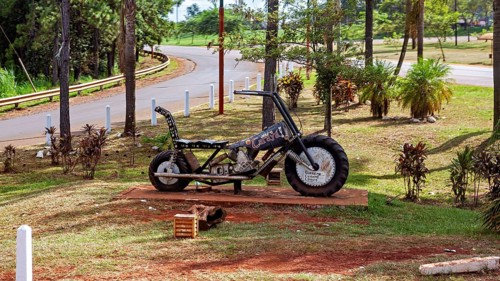 A metal motorbike bust stands in a park with green spaces and trees, placed on a pedestal, A