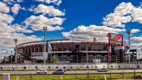 A modern stadium with stands and floodlights under a clear sky and passing cars, the River Plate