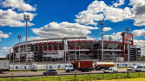 A large stadium with several stands and floodlights under a blue sky and passing vehicles, the