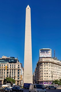 A tall obelisk stands between modern buildings under a clear blue sky, The Great Obelisk in the