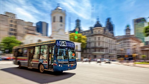 A colourful bus drives through a busy city with historic buildings and a blue sky in the
