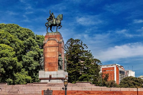 An equestrian monument on a pedestal surrounded by trees and buildings under a blue sky, The city