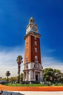 A tall brick clock tower next to palm trees under a clear blue sky, The city of Buenos Aires in