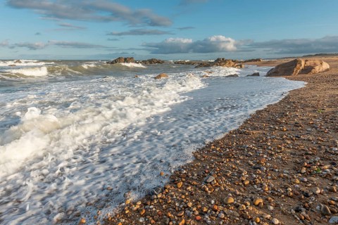 The waves crash gently against a rocky shoreline at sunset, creating a tranquil atmosphere. The