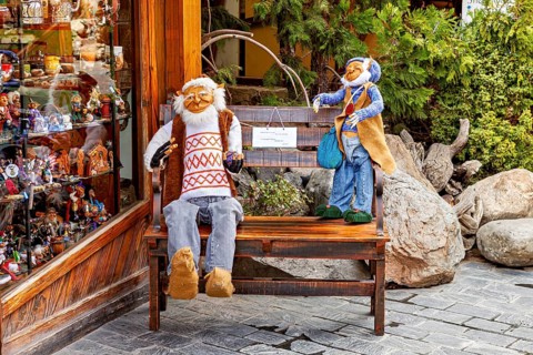 Two dolls sit on a bench in front of a shop window, surrounded by plants and wooden surfaces,