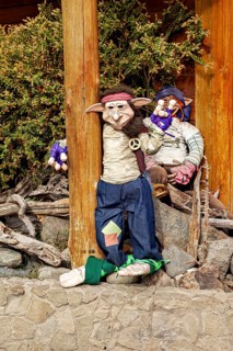 Two dolls lean against tree trunks next to a stone wall, decorated in a rustic setting, dwarves and