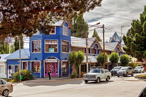 A blue, two-storey building in a busy street with parked cars and trees, street scene in El Chalten