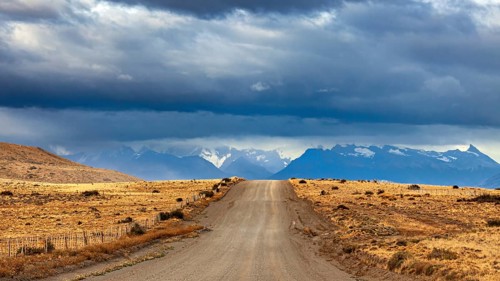 A long gravel road leads to a distant mountain range under a cloudy sky, The Patagonian landscape