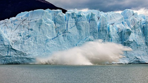 A glacier collapses into the water, blocks of ice tumble down. Threatening clouds add to the