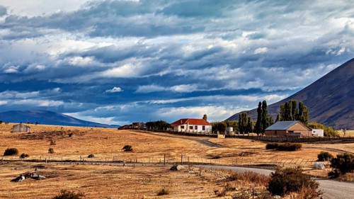 Lonely house in a rural setting with fields and mountains under a dramatic sky, The Patagonian