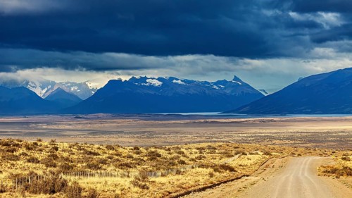 A gravel road leads to a mountain range under a dramatic cloudy sky, The Patagonian landscape near