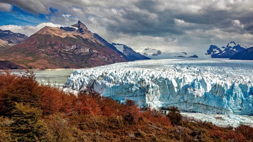 Imposing glacier front in a majestic mountain landscape with autumnal trees and dramatic skies, The