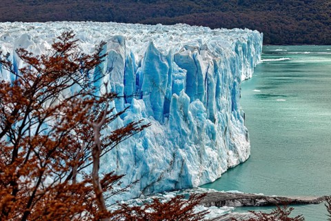 The glacier ends at an edge above a turquoise-coloured body of water, framed by autumnal trees, The