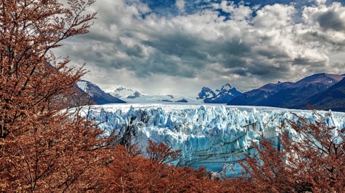 Blue glacier in the middle of a mountainous landscape with dramatic clouds and autumn leaves, The