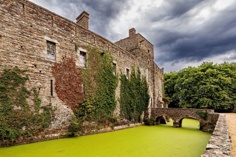 Side view of a castle with a bridge over a moss-covered moat under a cloudy sky, Pirou Castle in