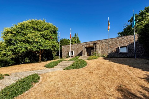 A modern building with flags, surrounded by trees and a dry meadow under a clear sky, German