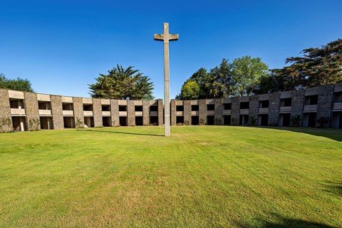 A stone cross sits enthroned in the centre of a green, circular courtyard surrounded by a concrete