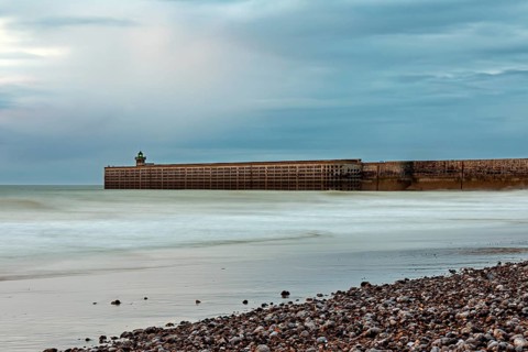 A breakwater juts into the sea, surrounded by a pebble beach and a cloudy sky, The pier and beach