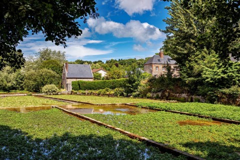 A sprawling green garden with water features and a house on the horizon under a blue sky, the