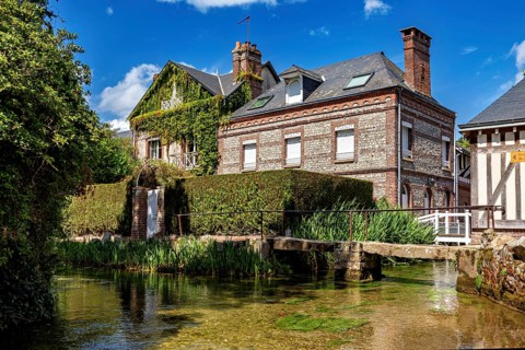 A brick house covered with climbing plants next to a clear body of water and lush vegetation, The