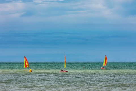 Three boats with yellow sails on the sea under a cloudy sky, surfers on the sea in Normandy, France