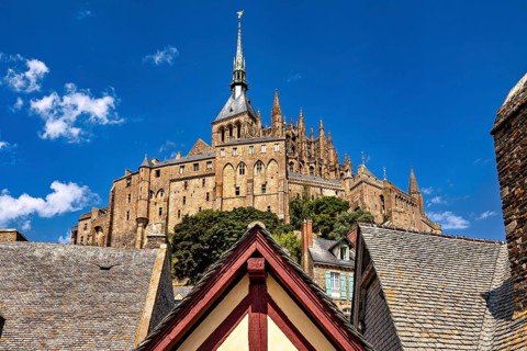 Medieval castle with a towering spire and rooftops against a clear blue sky, Mont Saint Michel in