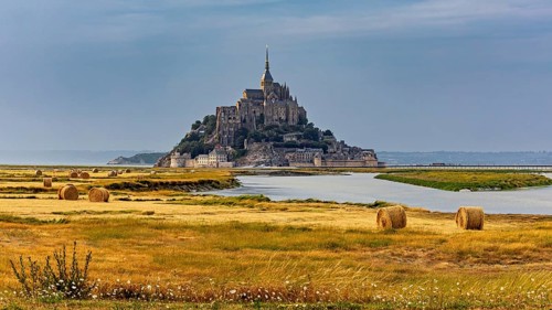 A castle rises in the middle of fields and a river, surrounded by bales of straw, Mont Saint Michel