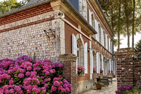Stone house with red bricks and white shutters surrounded by flowering hydrangeas and cobbled path,