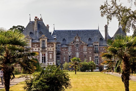A historic castle with towers, surrounded by gardens and palm trees, on a sunny day,