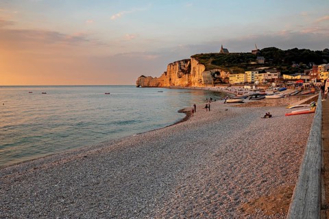 Beach with pebbles and boats, surrounded by rocks and a village, in the warm light of the sunset,