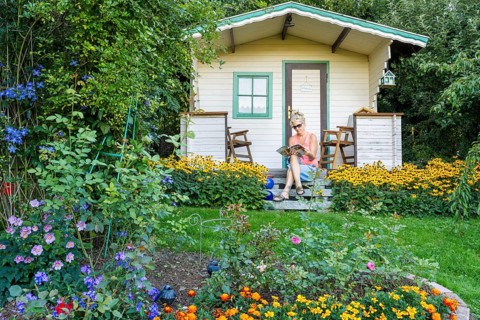 A woman sits relaxed on the small veranda of a garden shed and reads. Blooming summer flowers in